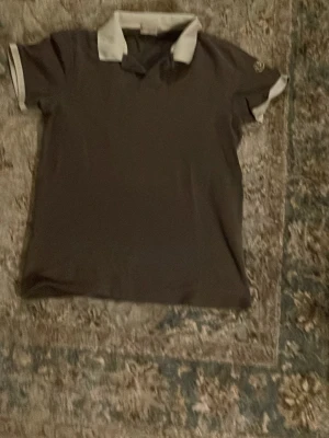 Monclear piké  - Snygg brun pikétröja med vit krage och vita detaljer på ärmsluten. Klassisk passform och kortärmad modell, perfekt till många olika outfits. Liten logga på ärmen för extra stil från Monclear kostar typ 3000kr ny pris