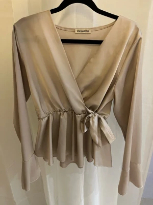 Beige omlottblus med knytband - Stilren beige omlottblus från New Collection med långa ärmar och volangkant nedtill. Blusen har ett knytband i midjan och djup v-ringning.