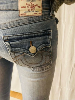 Ljusblå True Religion True Religion Joey Low Rise Flare-jeans  - Säljer ett par ljusblå jeans från True Religion  True Religion Joey Low Rise Flare-jeans med ikoniska bakfickor och snygga kontrastsömmar. Modellen har bootcut och klassisk jeanslook med knappdetalj på fickan. Perfekta för dig som vill ha en avslappnad men trendig stil.