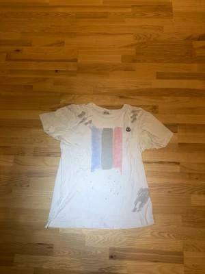 Vit t-shirt med tryck från Moncler - Vit t-shirt från Moncler med korta ärmar och tryck i blått, grått och rött framtill. Liten Moncler-logga på bröstet och unika grå detaljer på axel och nederkant. Perfekt för dig som gillar streetwear och vill sticka ut.