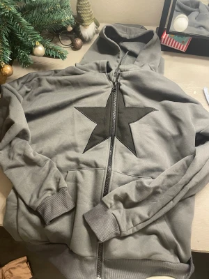 Grå hoodie med svart stjärna - Snygg grå hoodie med dragkedja och stor svart stjärna på bröstet. Tröjan har huva och ribbade muddar vid ärmslut och nederkant. Perfekt för en avslappnad och cool stil.