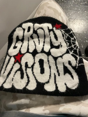 Svart mössa Groty Visions tryck - Cool svart mössa med vit text 'Groty Visions' i stor, grafisk stil och röda stjärnor. Ena sidan har ett vitt spindelnätsmönster. Perfekt för dig som gillar streetwear och unika accessoarer.