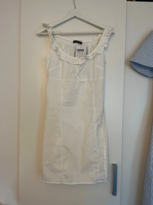 Vit klänning med volang från Brandy Melville - Helt oanvänd vit klänning från Brandy Melville med volangdetaljer vid axelbanden och knappar framtill. Klänningen är ärmlös och har en rak passform samt band man kan knyta antingen framtill eller på ryggen. Den är liten i storleken därför skulle jag rekommendera dig som bär storlek 36, och inte 38 som står på plagget.