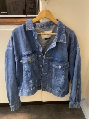 Blå jeansjacka från H&M, storlek M - Klassisk blå jeansjacka från H&M i storlek M. Jackan har en avslappnad passform, två bröstfickor med lock och knappar framtill. Perfekt att slänga över en hoodie eller t-shirt för en cool vardagslook.
