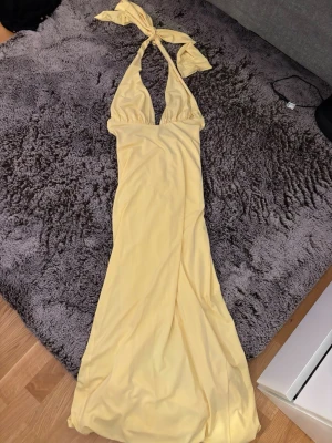 Gul långklänning med knyt i nacken - Snygg gul långklänning med djup v-ringning och knyt i nacken. Klänningen har en figurnära passform och är ärmlös, perfekt för sommarens alla tillfällen. Tillverkad i ett mjukt och följsamt material som ger en skön känsla. Använd 1 gång. Köpt för 1200 kr 