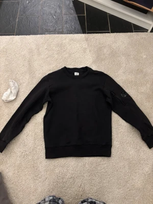 Svart sweatshirt från C.P. Company - Svart sweatshirt med rund hals från C.P. Company. Tröjan har en ikonisk linsdetalj på ena ärmen och ribbade muddar vid ärmslut och nederkant. Perfekt för en stilren och avslappnad look.