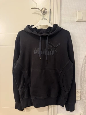 Svart hoodie från Puma - Svart hoodie från Puma med broderad logga och puma på bröstet. Klassisk modell med huva och dragsnören samt känguruficka framtill. Perfekt för en avslappnad och sportig stil.