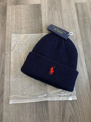 Mörkblå ribbstickad mössa Polo - Mörkblå ribbstickad mössa från Polo Ralph Lauren med klassisk uppvikt kant och röd broderad logga framtill. Perfekt accessoar för kyliga dagar och enkel att matcha med olika outfits.