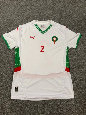 Marocko fotbollströja Puma Hakimi #2 - Säljer en vit fotbollströja från Puma med Marockos landslagsemblem, rött nummer 2 och Hakimi på ryggen. Tröjan har gröna och röda detaljer på ärmar och sidor samt korta ärmar och v-ringning. Perfekt för dig som älskar fotboll och vill stötta Marocko. Står XL men känns som M