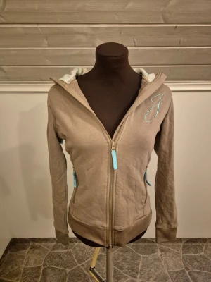 Zipup hoodie med blommig huva - Supersöt beige zipup hoodie med dragkedja och turkosa detaljer. Huvan har ett blommigt foder i vitt och turkost, och det finns en broderad J på bröstet🌸🩵 Köpt i en djuraffär så är egentligen en tröja för ridning men man kan ha den närsom✨️ Står m men passar mer s. Ena änden på bandet i huvan har tappt sin lilla plastgrej så där är den knuten men är annars som nyskick. Det är bara att skriva om man har frågor!😊💖