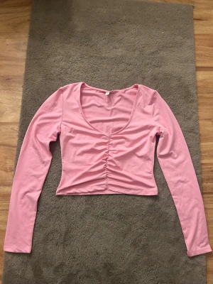 Rosa croppad topp med rynk framtill - Säljer en ljusrosa croppad topp med långa ärmar och snygg rynkdetalj framtill. Toppen har en djup rundringning och är gjord i mjuk bomullsmix som sitter skönt på kroppen. Perfekt för dig som vill ha en enkel men trendig look.