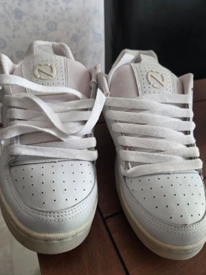 Vita sneakers med perforerad tå - Snygga vita sneakers med perforerad tå och tjocka vita snören. Skorna har en låg profil och en stilren design med diskret logga på plösen. Ovandelen är i syntetmaterial och mesh för bra andningsförmåga. Perfekta för en clean och fräsch look.Fläckarna i skorna går säkert att tvätta bort.