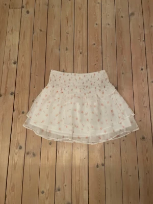 Vit blommig volangkjol - Supersöt kort kjol i vitt med små rosa blommor och volangdetaljer. Kjolen har en bred, smockad midja som ger en bekväm passform och ett luftigt, lager-på-lager-utseende. Perfekt för vår och sommar!