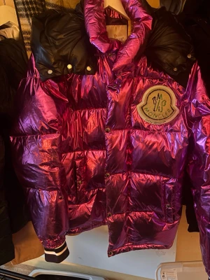 Moncler x Palm Angels metallic dunjacka - Snygg metallic rosa dunjacka från Moncler x Palm Angels med stor logopatch på bröstet och coolt innerfoder med Palm Angels-tryck. Jackan har svart axelparti, quiltad design, dragkedja och tryckknappar. Perfekt statement piece för vintern. Nypris 26000