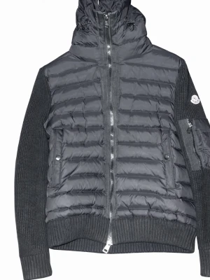 Svart dunjacka från Moncler - Snygg svart dunjacka från Moncler med quiltad front och rygg, stickade ärmar och muddar. Jackan har hög krage med dragsko, två sidofickor med knappar och dragkedja framtill. Perfekt för kalla dagar och riktigt stilren look.