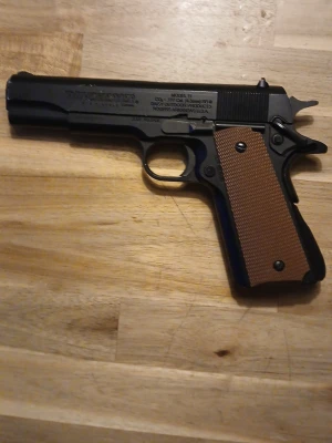 Svart airsoftpistol med brunt grepp - Säljer en svart airsoftpistol från Winchester med klassisk design och brunt räfflat grepp. Pistolen är tillverkad i metall och plast och har tydliga markeringar på sidan. Perfekt för airsoft eller samling.