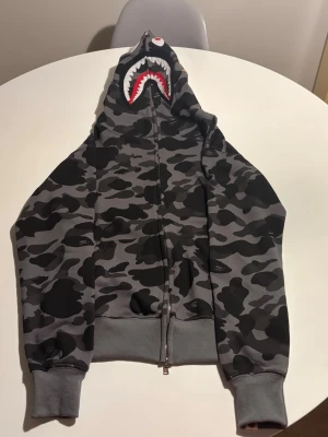 Grå camo hoodie från BAPE - Säljer en grå camo zip hoodie från BAPE med ikonisk hajdesign på luvan och broderade bokstäver i färgerna orange, blå och röd. Hoodien har ribbade muddar och är tillverkad i mjukt bomullsmaterial. Perfekt för dig som vill sticka ut med streetwear-stil.