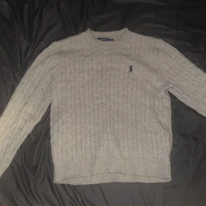 Grå kabelstickad tröja Polo Ralph Lauren - Kabelstickad grå tröja från Polo Ralph Lauren med klassisk rund halsringning och broderad logga på bröstet. Tröjan har ribbade muddar och är långärmad, perfekt för lager på lager under kyliga dagar. Sitter som storlek M 