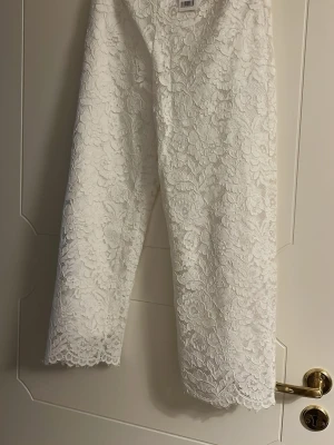 Vita spetsbyxor från Gina Tricot - Säljer ett par vita byxor från Gina Tricot med vackert blommigt spetsmönster. Byxorna har croppad längd och rak passform, perfekta för dig som vill ha något extra till outfiten. Supersnygga detaljer och riktigt trendig look! Längd-93 cm.