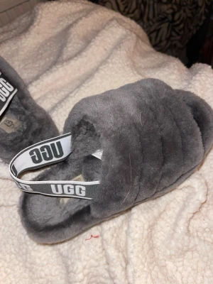 Grå fluffiga tofflor från UGG - Mysiga grå tofflor från UGG med fluffig päls och elastiskt band med UGG-logga. Perfekta att glida runt i hemma för en extra cozy känsla. Tofflorna har en rund tå och platt sula storlek 37.