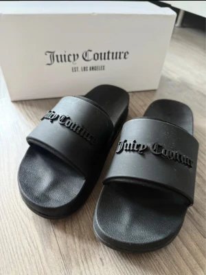 Svarta sandaler från Juicy Couture - Svarta sandaler från Juicy Couture med bred rem och upphöjd logga framtill. Sulan har ett stort Juicy Couture-mönster undertill. Tillverkade i syntetmaterial och har en platt sula för skön komfort. Perfekta för sommaren och riktigt snygga till alla outfits.