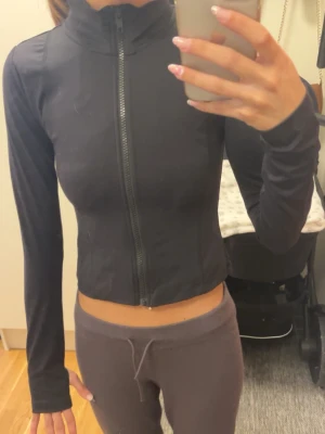 Svart tränings ziptröja - Säljer en svart croppad ziptröja med lång ärm och dragkedja framtill. Tröjan har en tajt passform och enkel, stilren design. Perfekt för dig som gillar en sportig och trendig look. Strlk 146-152