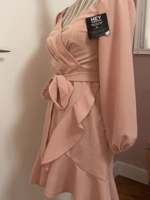 Helt oanvänd rosa klänning från Nelly  - Supersöt rosa klänning från Nelly med långa ärmar, volangdetaljer och knytning i midjan. Den är perfekt för vår och sommar. Materialet känns mjukt och lätt. Klänningen är oanvänd och har lapparna kvar. 