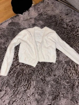 Vit fluffig stickad kofta - Mysig vit stickad kofta med öppen front och lång ärm. Koftan har en fluffig och mjuk känsla, perfekt att slänga på sig när det är kyligt. Enkel och stilren design som passar till många outfits.