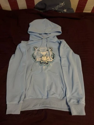 Ljusblå Kenzo hoodie med tigertryck - Snygg ljusblå hoodie från Kenzo med ikoniskt tigertryck och texten 'Kenzo Paris' på bröstet. Hoodien har huva med dragsko, magficka och ribbade muddar vid ärmar och nederkant. Perfekt för dig som vill ha en streetwear vibe och sticka ut lite extra. Hoodien är storlek M men sitter mer som Large/L. Hoodien har aldrig använts
