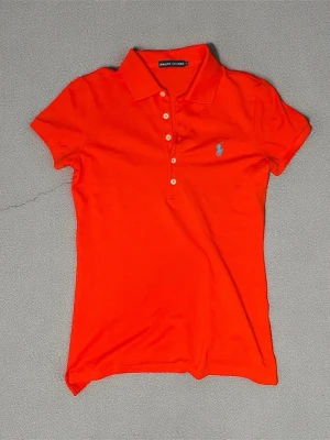 Pikétröja från Ralph Lauren, Stl S, i nytt skick.  - Snygg orange-röd Ralph Lauren pikétröja – stl S Fräsch och stilren piké från Ralph Lauren i en exklusiv orange-röd nyans. Klassisk modell med krage och fem knappar, samt den ikoniska ljusblå Polo-loggan broderad på bröstet för en elegant kontrast.  Tröjan är i nästintill nyskick och kommer från ett djur- och rökfritt hem. Perfekt för både vardag och finare tillfällen.  Storlek: S Skick: Nytt skick / mycket sparsamt använd