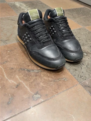 Valentino Garavani rockrunners svarta sneakers - Svarta Valentino rockrunner sneakers med klassiska nitar på sidorna och beige sula. Skorna har snörning och är tillverkade i skinn med detaljer i mocka. Perfekta för dig som gillar en stilren och edgy look. Mycket bra skick. Storlek 42 kan också passa 43.