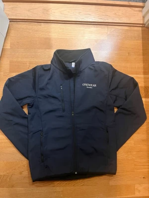 Svart vindjacka från ONEWear - Snygg svart vindjacka från ONEWear med hög krage och dragkedja framtill. Jackan har tre fickor med dragkedja och diskret logga på bröstet. Perfekt för blåsiga dagar och enkel att matcha med olika outfits.