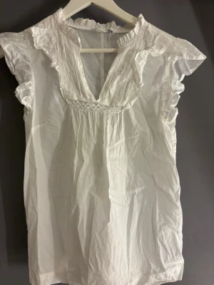 Vit volangblus från 157 XS - Söt vit blus från 157 i storlek XS med volangdetaljer vid halsringningen och ärmsluten. Blusen har en djup V-ringning och korta ärmar med rynkade kanter. Tillverkad i ett lätt och luftigt tyg, perfekt för en fräsch och stilren look.