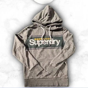 Grå hoodie från Superdry - Snygg grå hoodie från Superdry med stor logga och japansk text på bröstet. Hoodien har vit dragsko i huvan och en klassisk känguruficka framtill. Perfekt för en avslappnad streetstyle-look. Passar någon 170-185 
