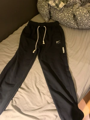 Svarta Nike mjukisbyxor med snörning - Svarta mjukisbyxor från Nike med vit snörning i midjan och elastisk linning. Byxorna har fickor med dragkedja och Nike-logga på ena benet. Perfekta för chill eller träning, tillverkade i mjukt bomullsmaterial. Endast provad!! Ej andvända 