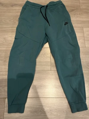 Blå mjukisbyxor från Nike - Snygga blå mjukisbyxor från Nike med svart snörning i midjan och logga på vänster ben. Byxorna har en relaxed passform och är gjorda i mjukt bomullsmaterial. Perfekta för chill eller träning, med ribbade muddar vid bensluten.