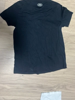 Svart t-shirt från Under Armour - Svart t-shirt från Under Armour med stor vit logga och text på bröstet. Passar 158/164, är skrynklig med stryks innan leverans.