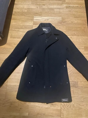 Peacoat / Kappa - Stilren svart kappa från Blend med klassisk krage och diskreta knappar framtill. Kappan har två fickor med knappdetaljer och dragkedja under knappslån. Perfekt för dig som gillar en enkel och tidlös look. Pris kan diskuteras.