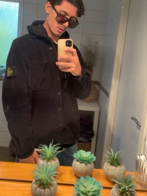 Svart overshirt från Stone Island - Säljer en svart overshirt från Stone Island i manchester med klassisk patch på vänster ärm. Skjortjackan har knäppning framtill, huva och lång ärm. Perfekt för lager-på-lager och en clean streetwear-look.