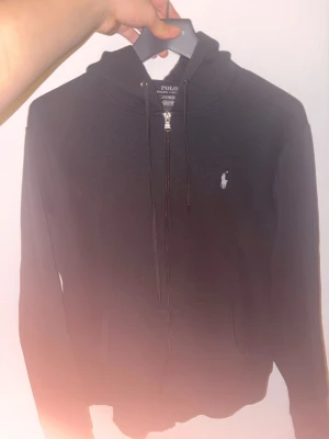 Svart hoodie från Polo Ralph Lauren - Klassisk svart hoodie från Polo Ralph Lauren med dragkedja framtill och vit broderad logga på bröstet. Hoodien har huva med dragsko och känguruficka. Tillverkad i mjukt bomullsmaterial som är skönt att bära.