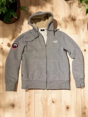 Grå zip hoodie från Superdry - Snygg grå hoodie från Superdry med dragkedja, mjukt teddyfoder i huvan och två fickor framtill. Broderad Superdry-logga på bröstet och en patch på ärmen. Perfekt för chill dagar och skolan