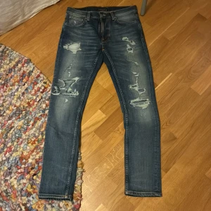 Blå slitna nudie jeans - Snygga blå jeans med slitningar och och hål vid knäna. Jeansen har klassisk femficksdesign och tydliga slitdetaljer framtill för en trendig look. Perfekta för dig som gillar en avslappnad och cool stil.