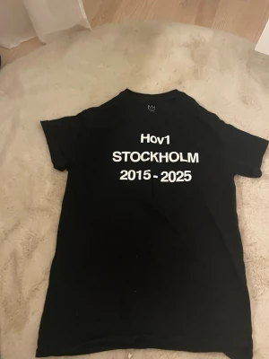 Svart Hov1 Stockholm t-shirt - Säljer en svart t-shirt från Hov1 med vit text fram och bak. På framsidan står det 'Hov1 STOCKHOLM 2015 - 2025' och på baksidan finns turnédatum och texten 'Vi Dansar Vidare I Livet'. Klassisk passform och rund hals. Perfekt för dig som gillar svensk musik. Den är från deras sista konsert och köpt för 700kr. Väldigt oversized! Använd typ en gång.