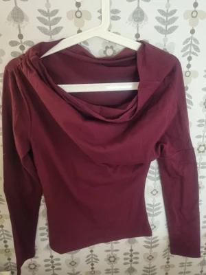 Vinröd långärmad topp med draperad krage - Säljer en vinröd off shoulder top med snyggt draperad krage. Toppen har en slim passform och är tillverkad i mjukt bomullsmaterial. Perfekt för dig som vill ha en stilren men ändå unik look.