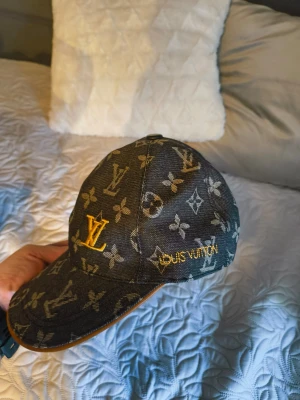  L V denimkeps monogram - Snygg keps  i svart denim med klassiskt monogrammönster i beige och gult. Broderad LV-logga framtill och text på sidan. Böjd skärm med gul kant och justerbar rem bak. Perfekt för dig som gillar exklusiv streetwear.