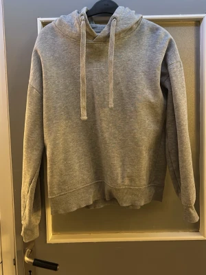 Grå basic hoodie med huva - En enkel och stilren grå hoodie med huva och dragsnören. Tillverkad i mjukt sweatshirtmaterial som är perfekt för chill dagar. Långärmad modell med ribbade muddar och avslut. Passar till det mesta och är ett måste i garderoben.