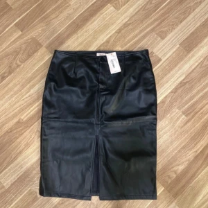 Svart pennkjol i skinnimitation från Nelly - Säljer en svart pennkjol från Nelly i skinnimitation med snygg slits framtill. Kjolen har en klassisk midjeform och stängs med knapp och dragkedja. Perfekt för dig som gillar en stilren och edgy look.