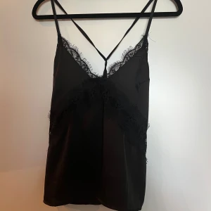 Svart spetslinne från NA-KD - Säljer ett svart linnetopp från NA-KD med tunna axelband och spetsdetaljer längs v-ringningen och över bröstet.