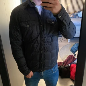 Mörkblå quiltad pufferjacka - Säljer en mörkblå pufferjacka med quiltad design och knappar framtill. Jackan har två bröstfickor med lock och knapp, samt klassisk krage. Passformen är normal och jackan är långärmad, perfekt för kyliga dagar.