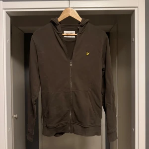Olivgrön hoodie från Lyle & Scott - Säljer en olivgrön hoodie från Lyle & Scott med dragkedja och huva. Tröjan har två fickor framtill och den klassiska gula loggan på bröstet. Perfekt för chill dagar eller när du vill ha en enkel och stilren look.